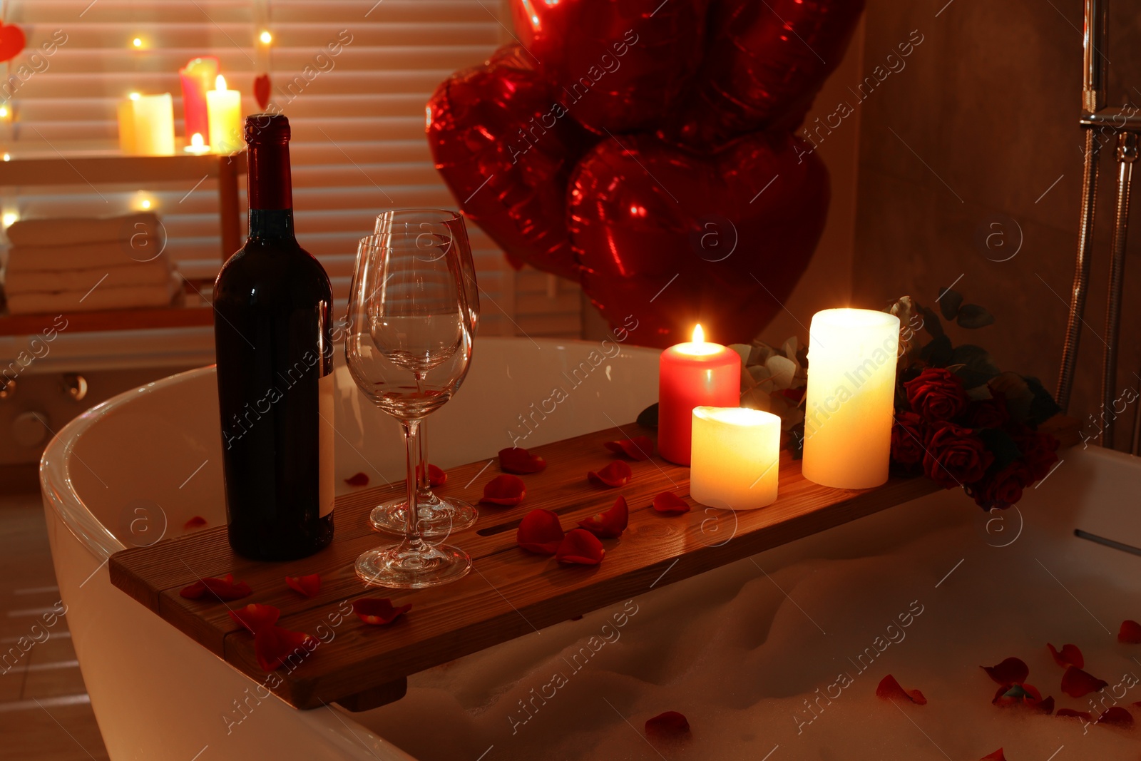 Photo of Wooden tray with wine, burning candles and rose petals on tub in bathroom. Valentine's day celebration