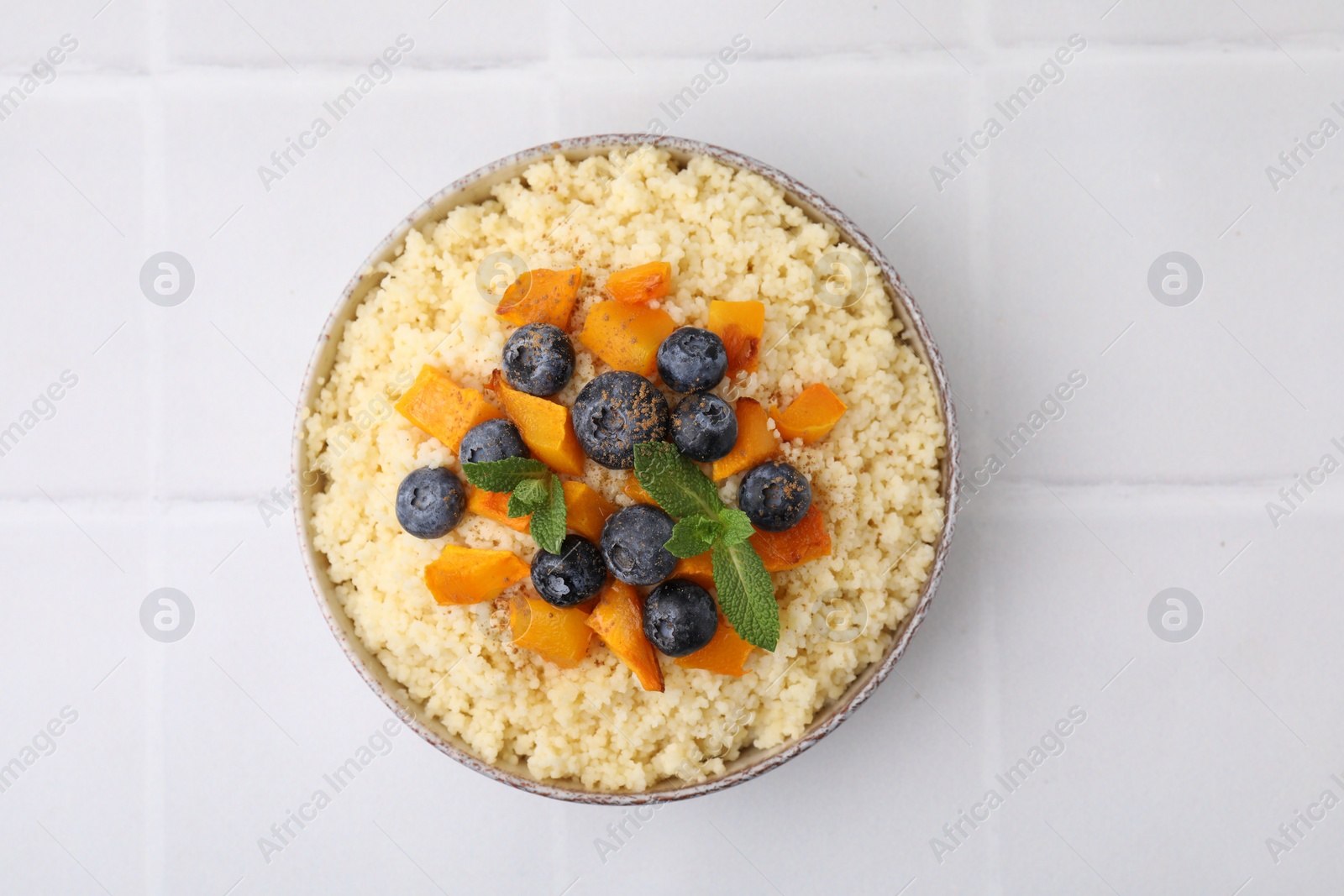 Photo of Bowl of tasty couscous with blueberries, pumpkin and mint on white tiled table, top view