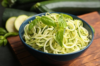 Photo of Delicious zucchini pasta with basil in bowl on wooden board, closeup