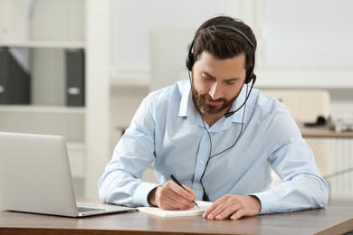 Photo of Hotline operator with headset writing something in notebook while working at wooden table