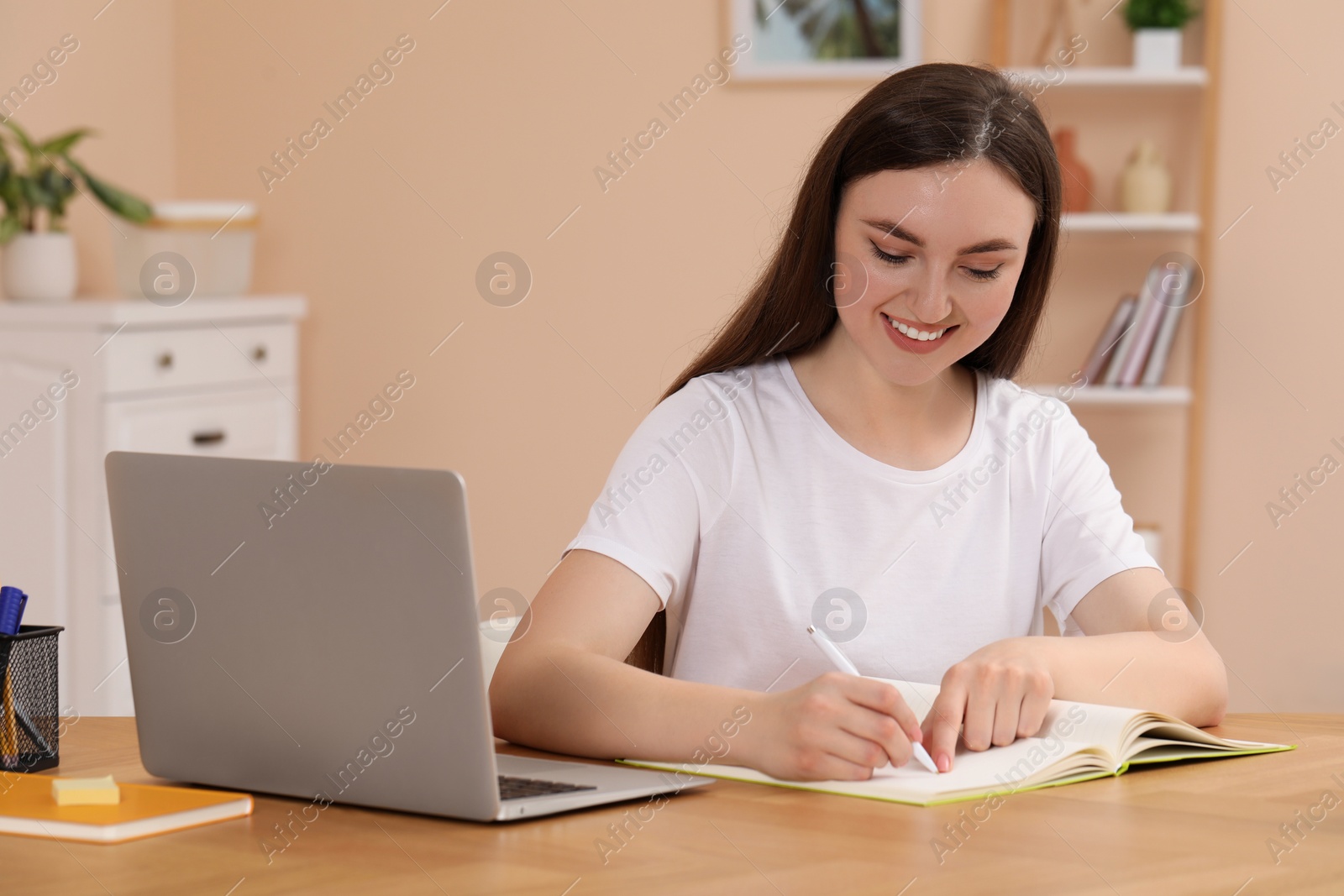 Photo of Woman writing in notebook while working on laptop at wooden table indoors