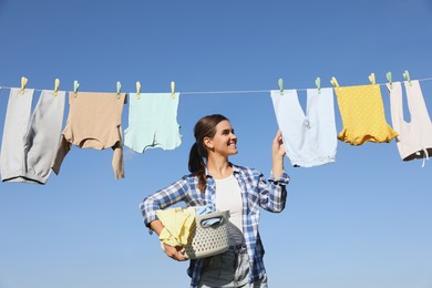 Smiling woman holding basket with baby clothes near washing line for drying under blue sky outdoors