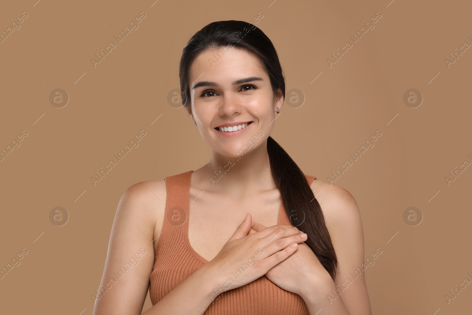 Photo of Thank you gesture. Beautiful grateful woman holding hands near heart on brown background