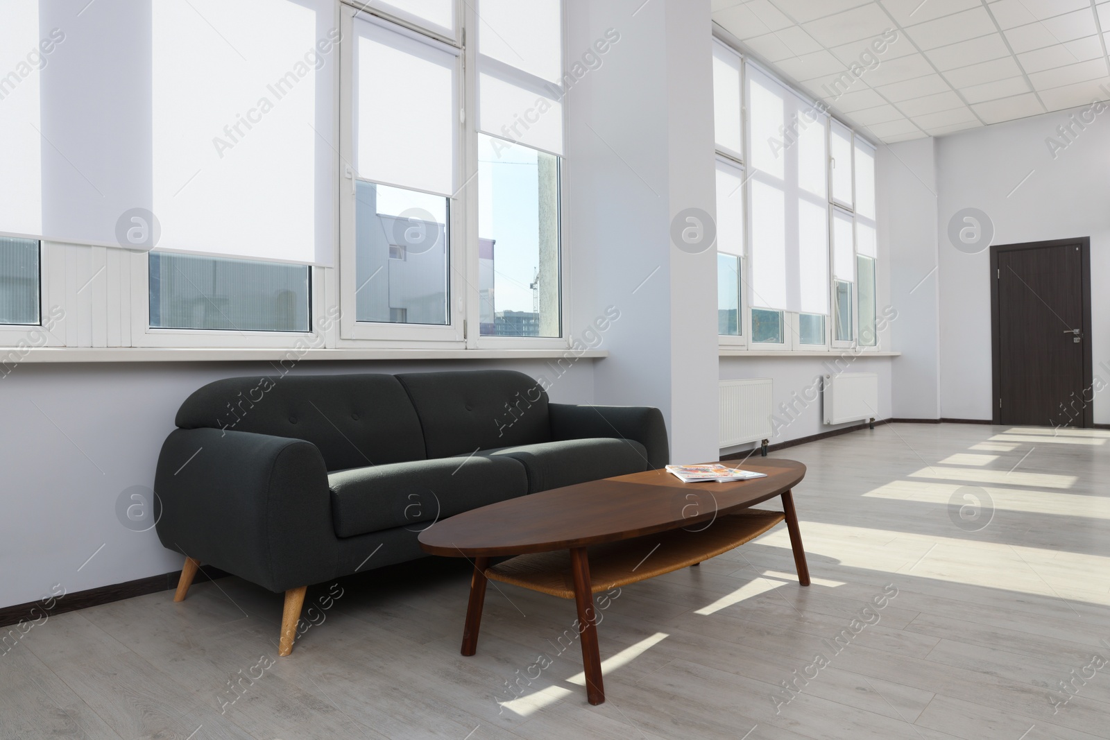 Photo of Comfortable sofa and table near large windows with white roller blinds indoors