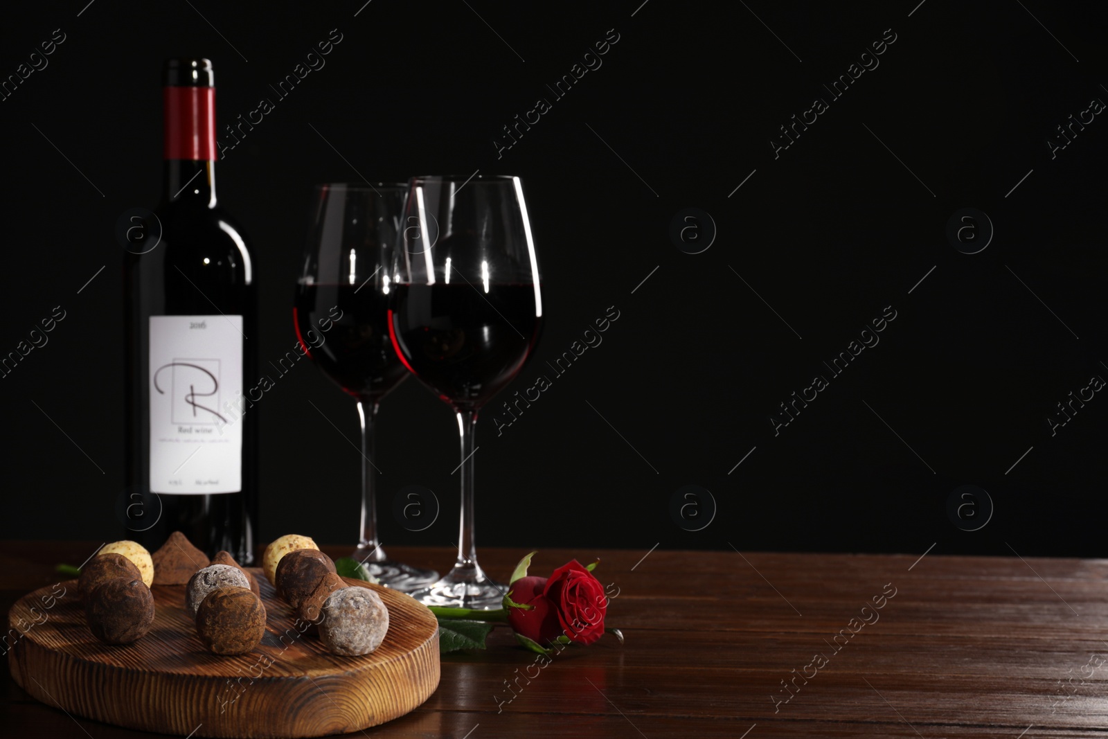 Photo of Red wine, chocolate truffles and rose flower on wooden table against dark background, space for text