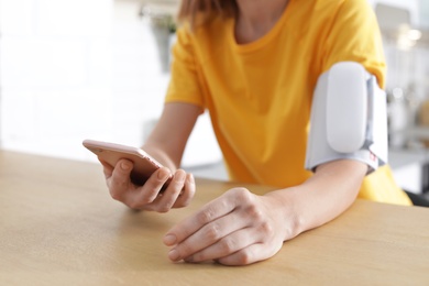 Woman checking blood pressure with modern monitor and smartphone at table indoors, closeup. Cardiology concept