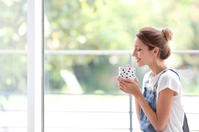 Photo of Young woman with cup of aromatic coffee near window at home