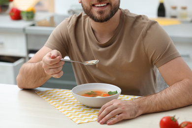 Young man eating tasty vegetable soup at table in kitchen, closeup