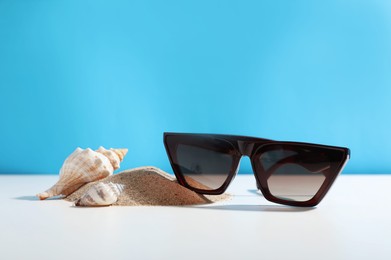 Photo of Stylish sunglasses, seashells and sand on white table against light blue background. Space for text