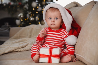 Photo of Cute baby in Santa hat with Christmas gift on sofa at home