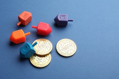Photo of Dreidels with Jewish letters and coins on blue background, space for text. Traditional Hanukkah game