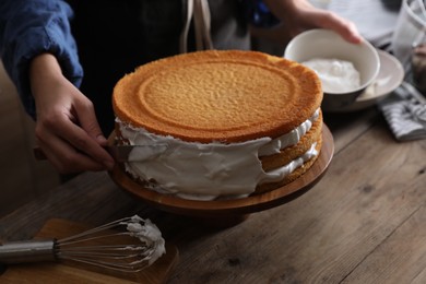 Woman smearing sides of sponge cake with cream at wooden table, closeup