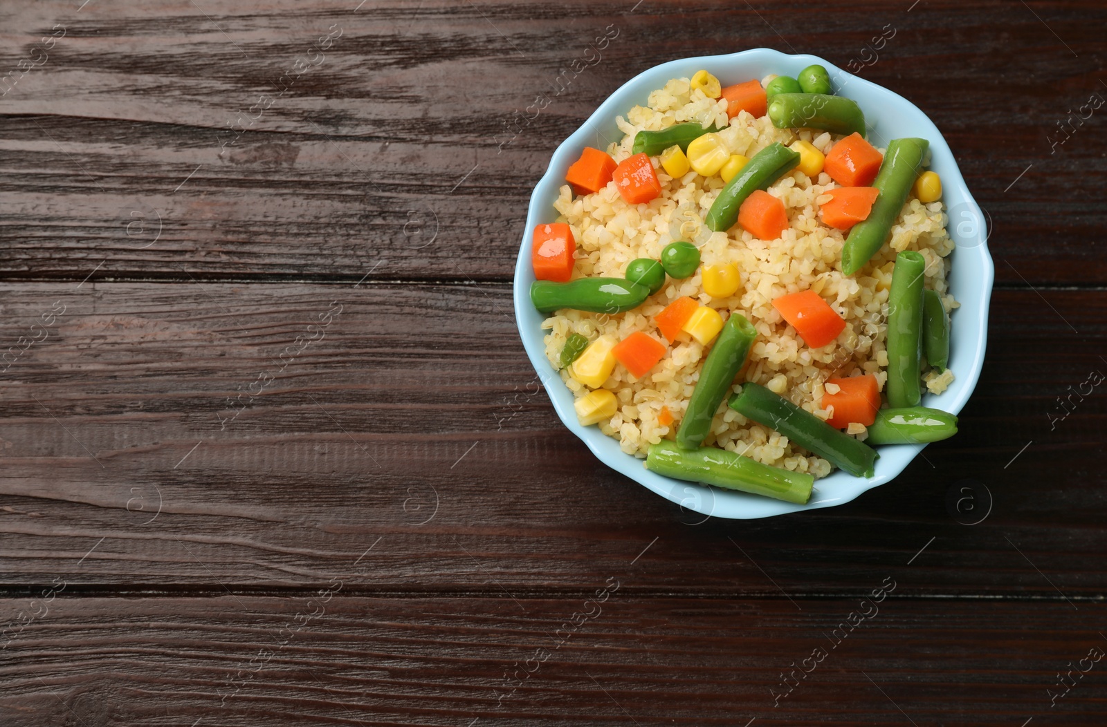 Photo of Delicious bulgur with vegetables in bowl on wooden table, top view. Space for text