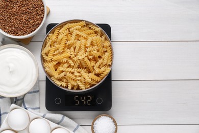 Photo of Electronic scales with uncooked pasta and different products on white wooden table, flat lay. Space for text