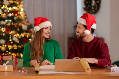 Photo of Happy couple in Santa hats decorating Christmas gift with wrapping paper at table in room
