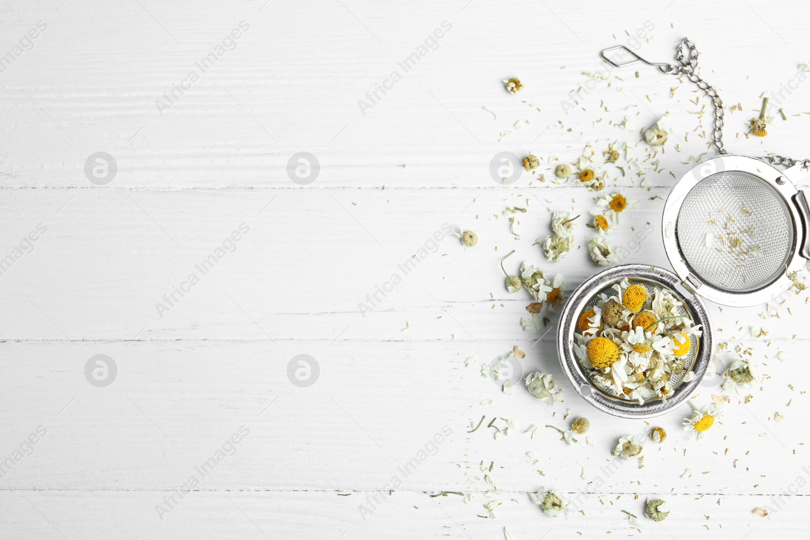 Photo of Dry chamomile flowers in infuser on white wooden table, flat lay. Space for text