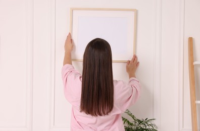 Woman hanging picture frame on white wall indoors, back view