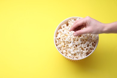 Photo of Woman taking delicious popcorn from paper bucket on yellow background, top view. Space for text