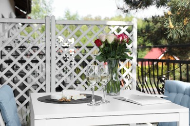 Photo of Vase with roses, open book, glasses of wine and snacks on white table at balcony