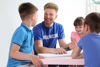 Photo of Young volunteer reading book with children at table indoors