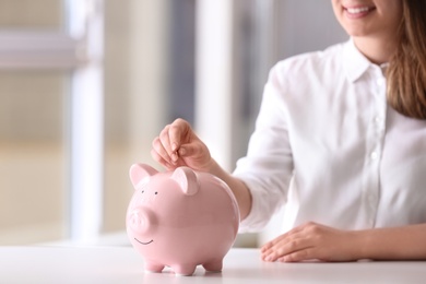 Woman putting coin into piggy bank at table indoors, closeup. Space for text