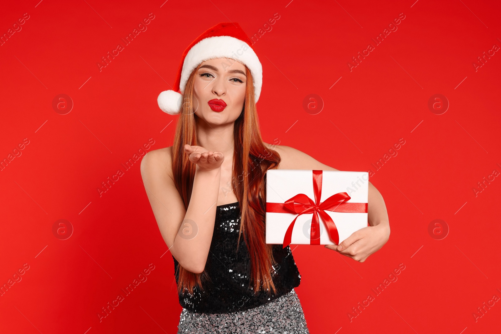 Photo of Young woman in Santa hat with Christmas gift blowing kiss on red background