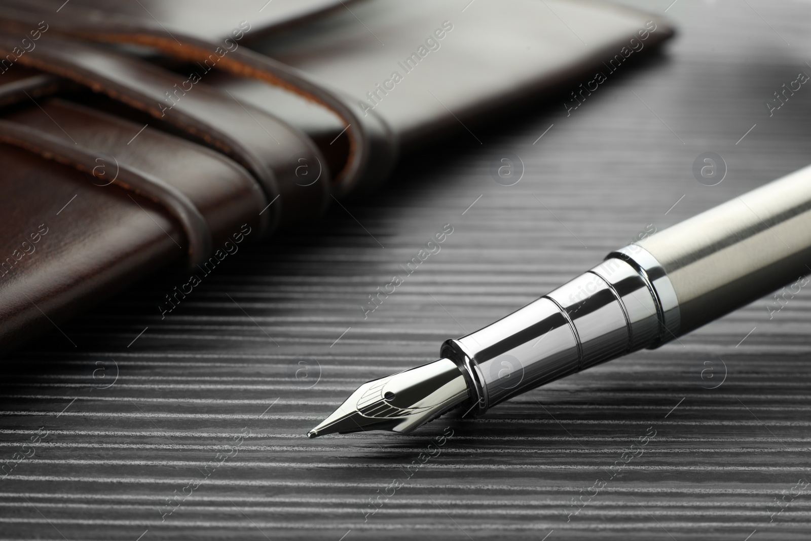 Photo of Stylish silver fountain pen and leather notebook on black wooden table, closeup