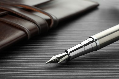 Stylish silver fountain pen and leather notebook on black wooden table, closeup