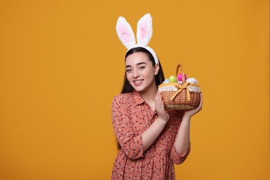 Photo of Happy woman in bunny ears headband holding wicker basket of painted Easter eggs on orange background. Space for text