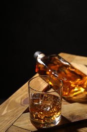 Whiskey with ice cubes in glass and bottle on wooden crate against black background, closeup
