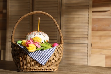 Photo of Traditional Easter cake with burning candle, dyed eggs and flowers in basket on wooden table indoors. Space for text