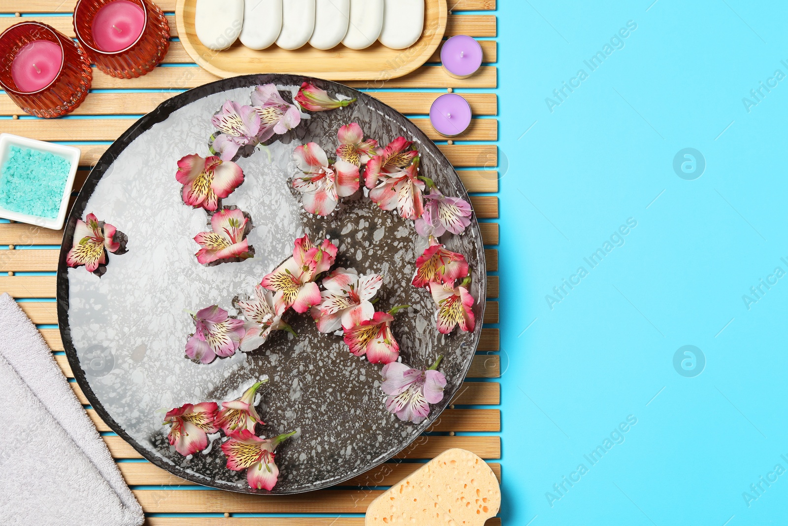 Photo of Bowl of water with flowers and different spa supplies on light blue background, flat lay. Space for text