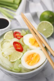 Photo of Bowl of delicious rice noodle soup with celery and egg on white marble table, closeup