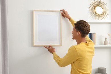 Photo of Young man hanging picture frame on white wall indoors, back view