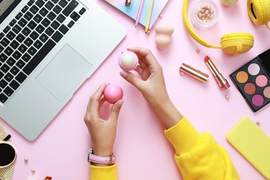 Photo of Young beauty blogger with lip balm near laptop and accessories on light pink background, top view