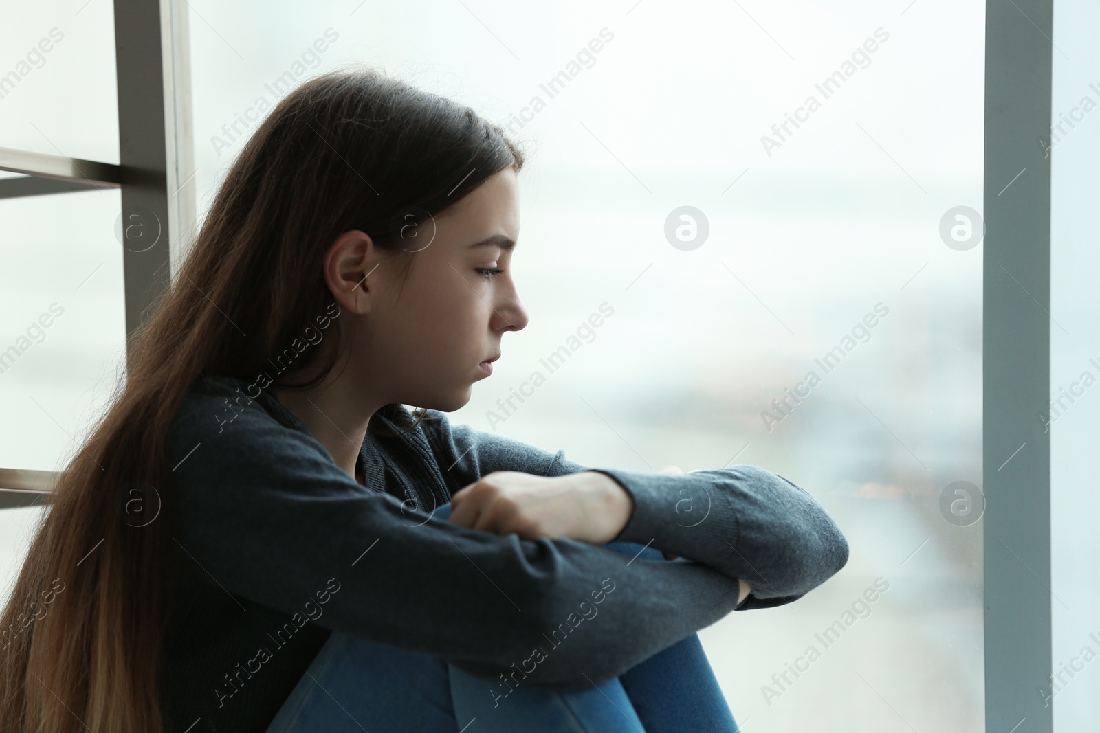 Photo of Upset teenage girl sitting at window indoors. Space for text