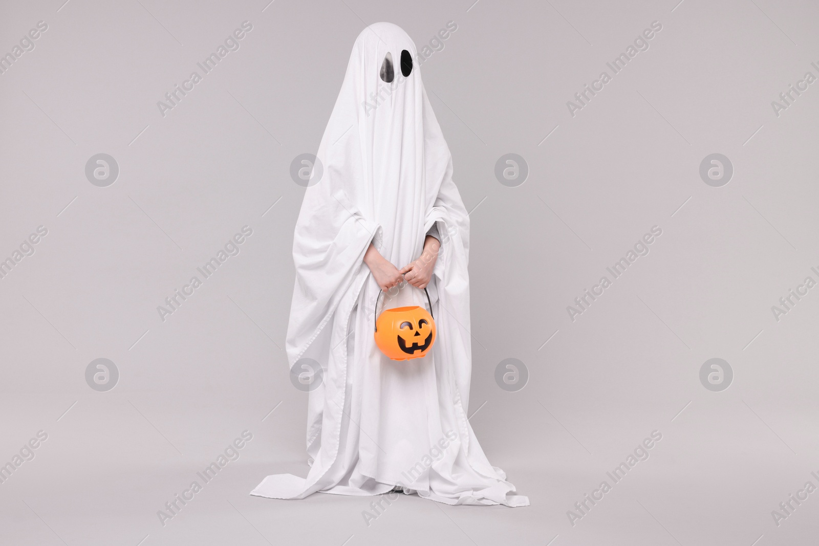 Photo of Child in white ghost costume holding pumpkin bucket on light grey background. Halloween celebration