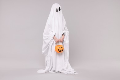 Child in white ghost costume holding pumpkin bucket on light grey background. Halloween celebration