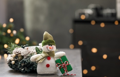 Photo of Snowman toy and Christmas decor on grey table against blurred festive lights. Space for text