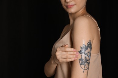 Photo of Woman applying cream onto her arm with tattoo on dark background, closeup. Space for text