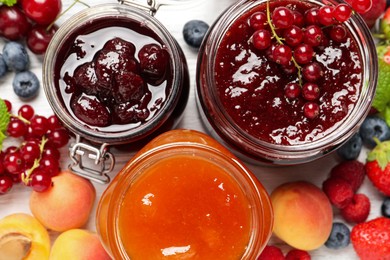 Jars with different jams and fresh fruits on white wooden table, flat lay