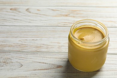 Spicy mustard in glass jar on white wooden table, space for text