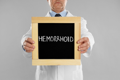 Photo of Doctor holding blackboard with word HEMORRHOID on light grey background, closeup