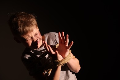 Scarred little boy with bruises tied up and taken hostage on dark background. Space for text
