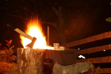 Photo of Tree stump with axe and burning firewood in metal brazier outdoors at night
