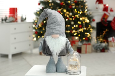 Photo of Funny Christmas gnome and magical snow globe on coffee table in room with festive decoration