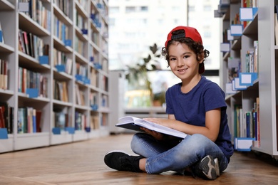 Photo of Cute little boy reading book on floor in library