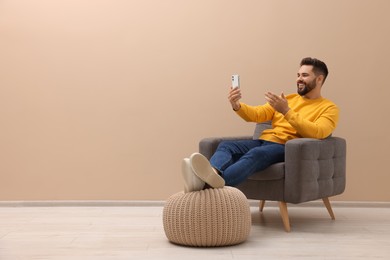 Photo of Handsome man having video chat via smartphone while sitting in armchair near beige wall indoors, space for text