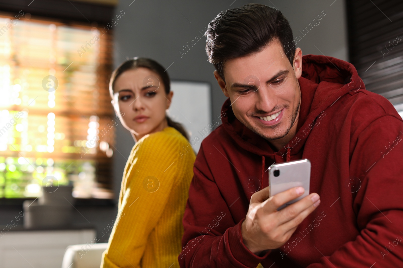 Photo of Distrustful woman peering into boyfriend's smartphone at home. Jealousy in relationship