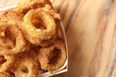 Cardboard box with crunchy fried onion rings on wooden background, closeup. Space for text